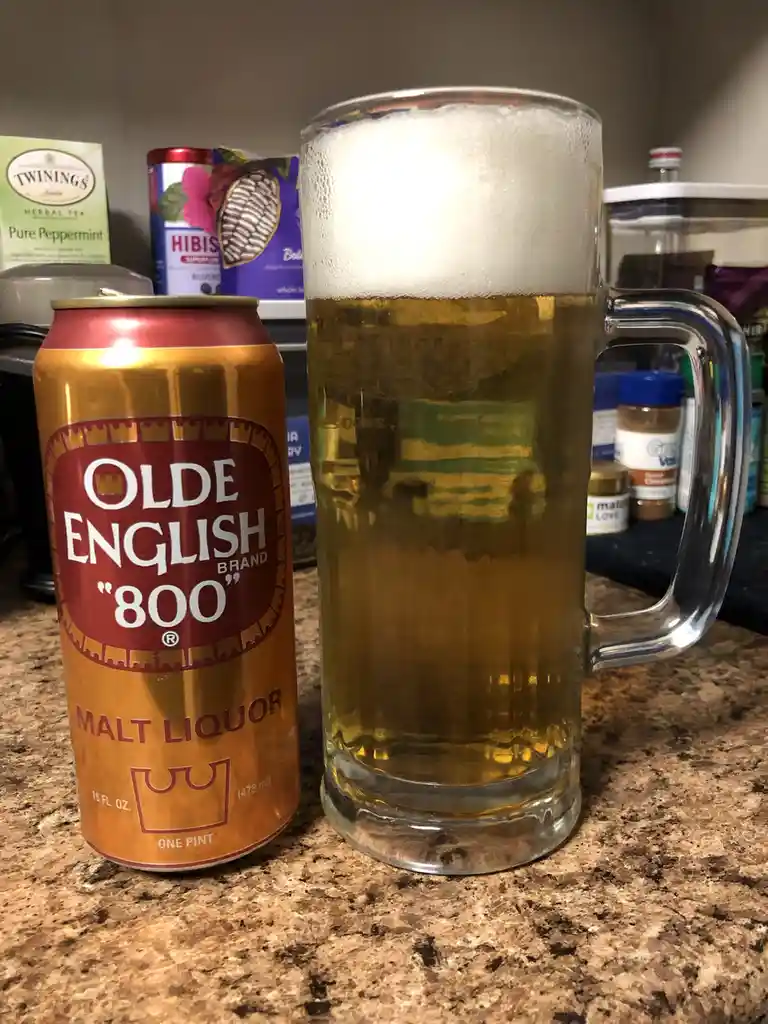 A can of Olde English 800 malt liquor sits next to a tall glass mug filled with beer and topped with foam, both resting on a…