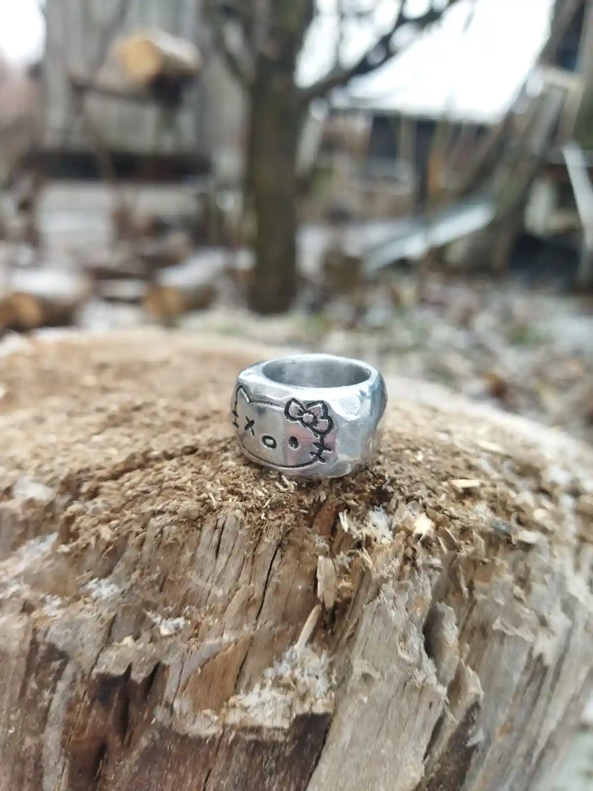 A silver ring with a Hello Kitty face design, featuring a bow and X's for eyes, sits on a rough, weathered tree stump…