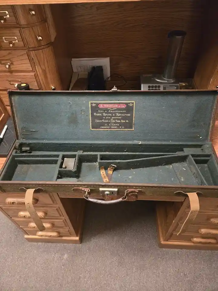 An old, empty, green-lined gun case with a brass plaque reading 'E. WHISTLER & Co. NEW & SECONDHAND GUNS, RIFLES, &…