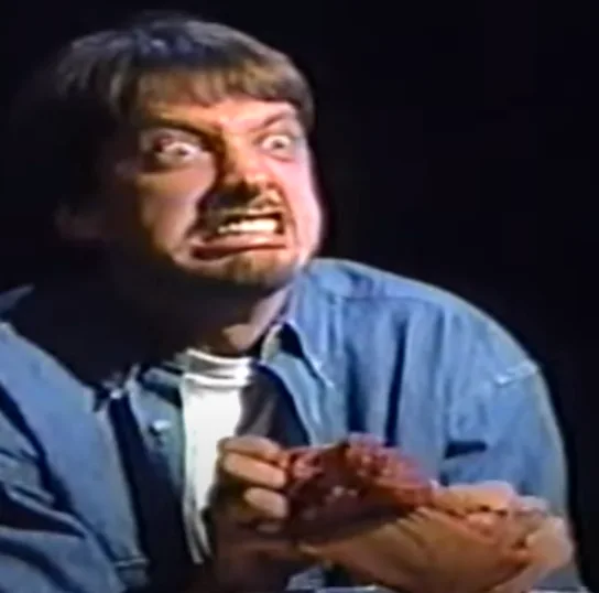 A man with a shocked, disgusted facial expression, wearing a blue denim shirt over a white t-shirt, holds a piece of…