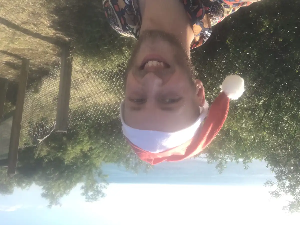 A man with a beard wearing a red and white Santa hat smiles at the camera while lying upside down outdoors. The background…