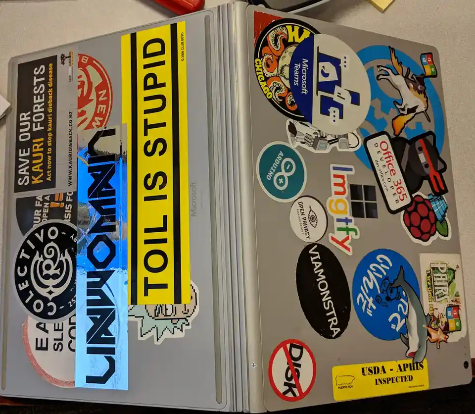 A silver laptop covered in numerous colorful stickers, including Microsoft Teams, Arduino, Raspberry Pi, and 'TOIL IS…