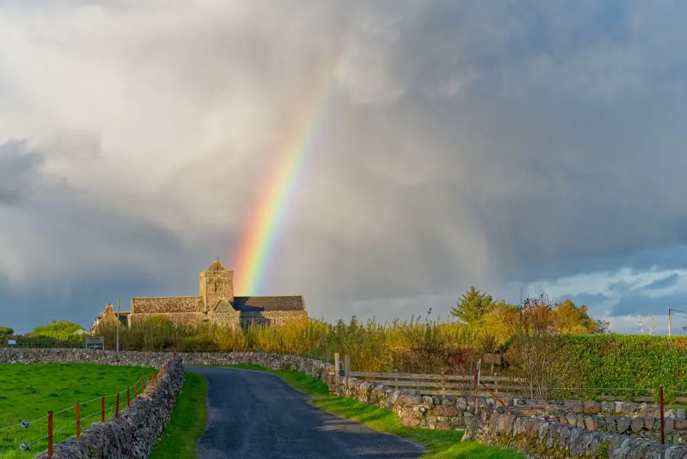 A scenic rural road leads to an old stone church under a dramatic sky with dark clouds. A vivid rainbow arcs from the left…