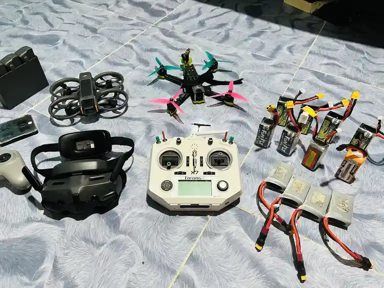 An overhead photo displays a collection of drone-related gear laid out on a textured white carpet, including two drones (one…