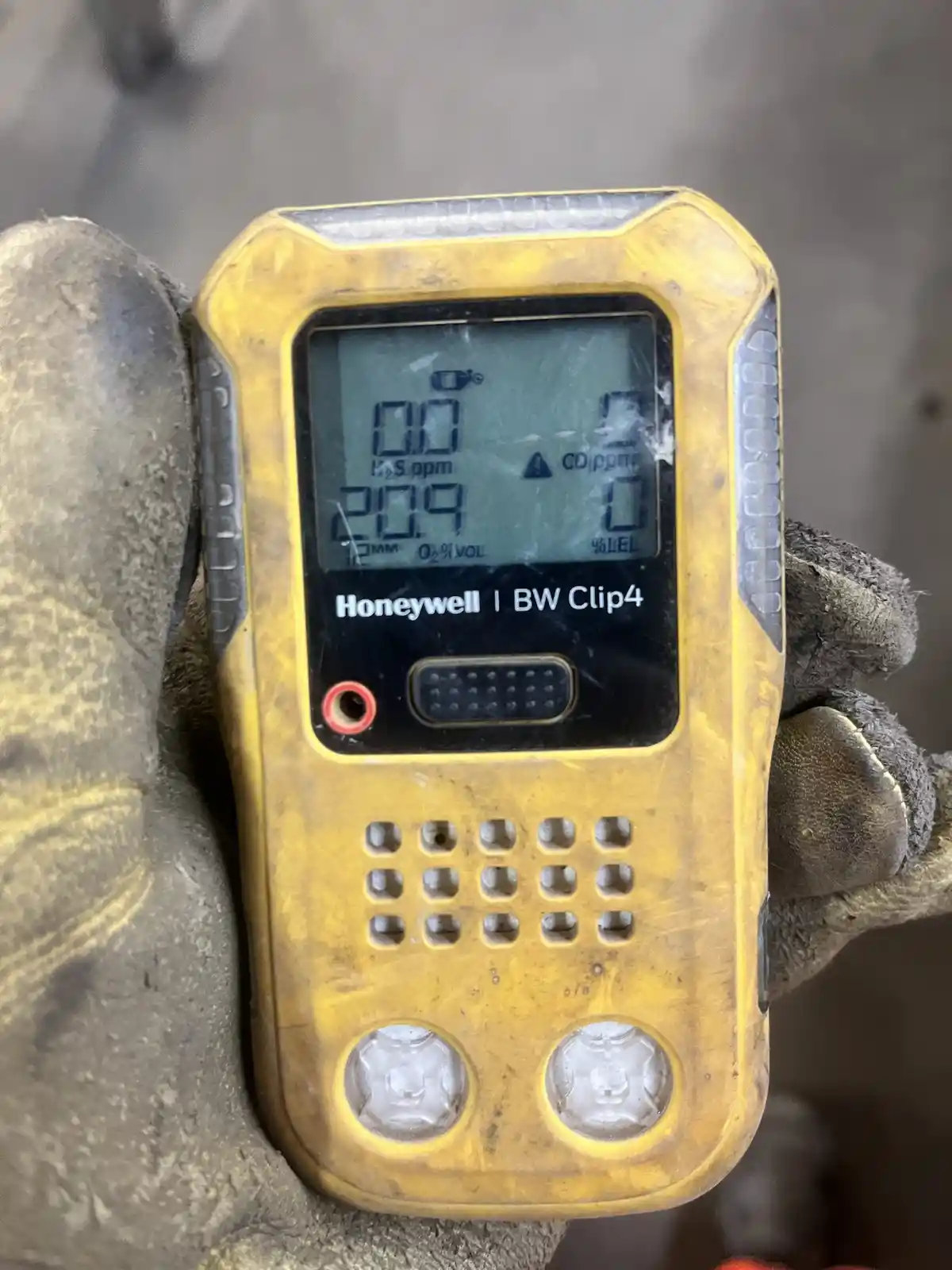 A close-up of a yellow Honeywell BW Clip4 gas detector held in a dirty work glove, showing a screen with readings: H2S at…