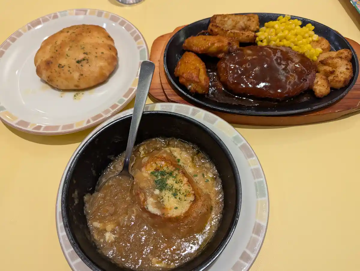 A table displays three dishes: a bowl of French onion soup with a spoon, a plate with a round bread roll, and a sizzling…