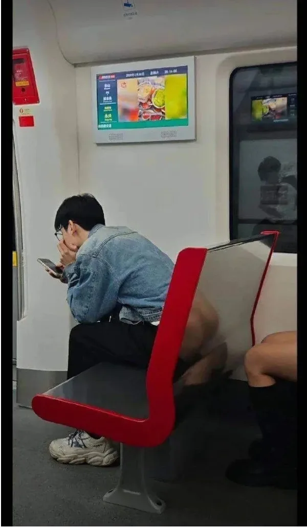 A person sits on a red train seat, hunched over their phone with hands covering their face, while their legs are visibly…