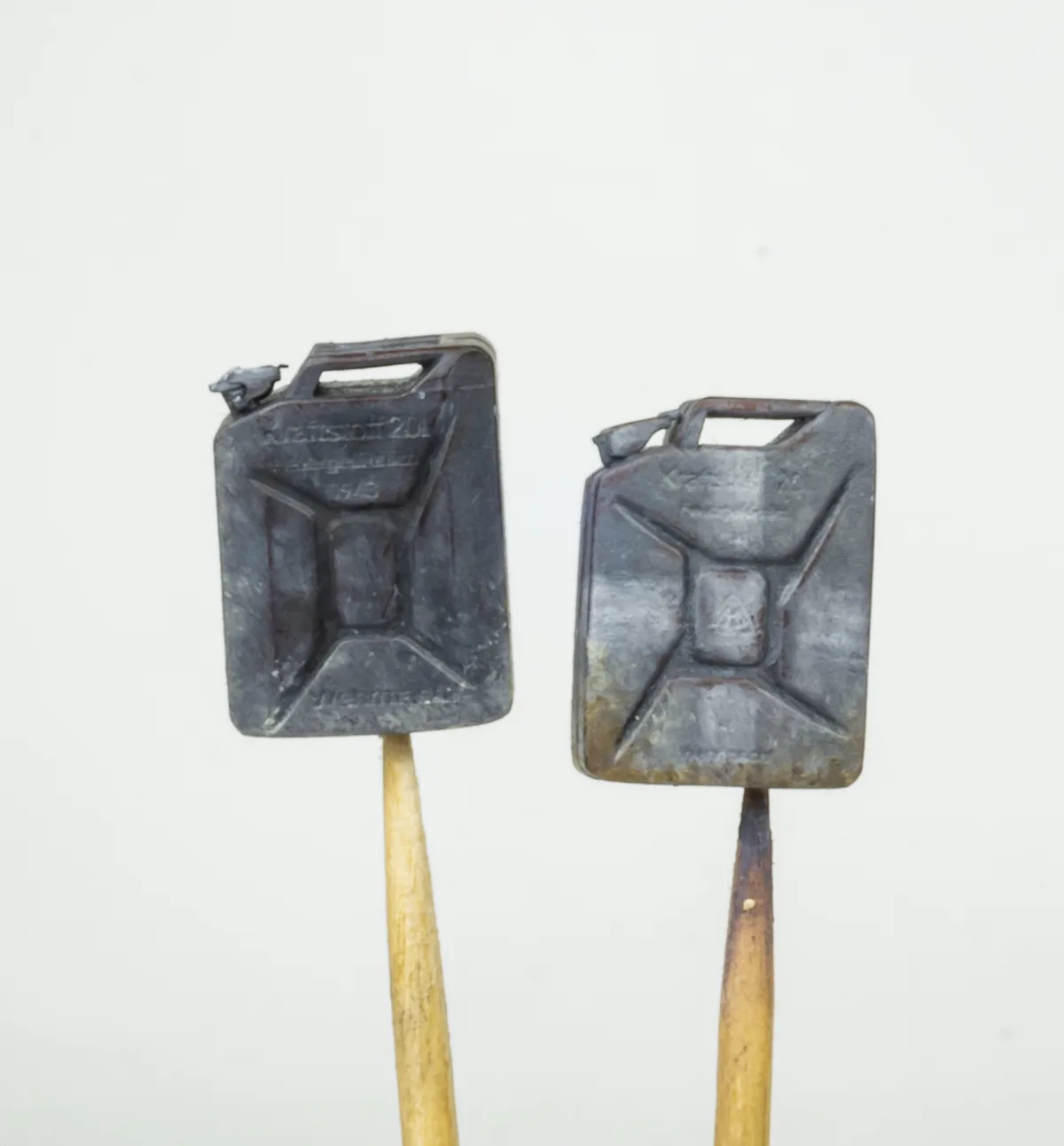 Two small, weathered metal fuel canisters, each mounted on a wooden toothpick against a plain white background. The…
