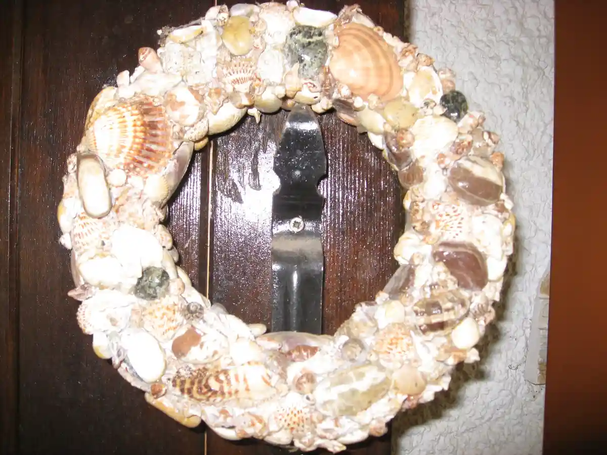 A circular wreath made of assorted seashells and small stones is mounted on a dark wooden door, surrounding a black metal…