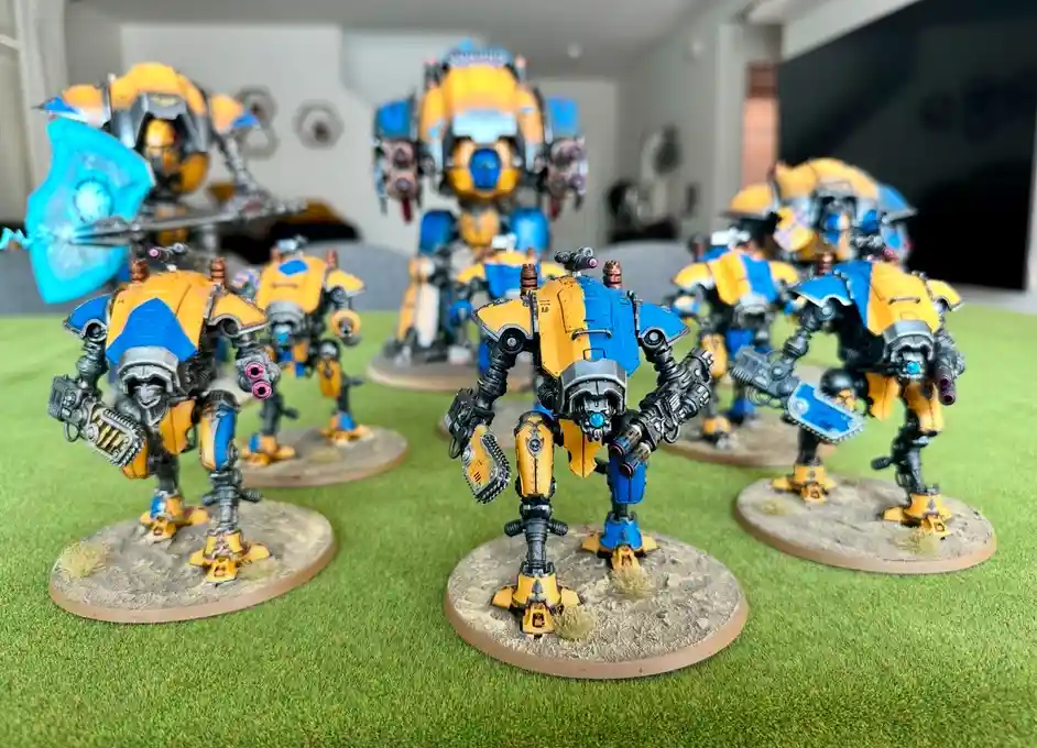 A photo of multiple yellow and blue painted miniature robotic figures standing on circular bases on green artificial grass…