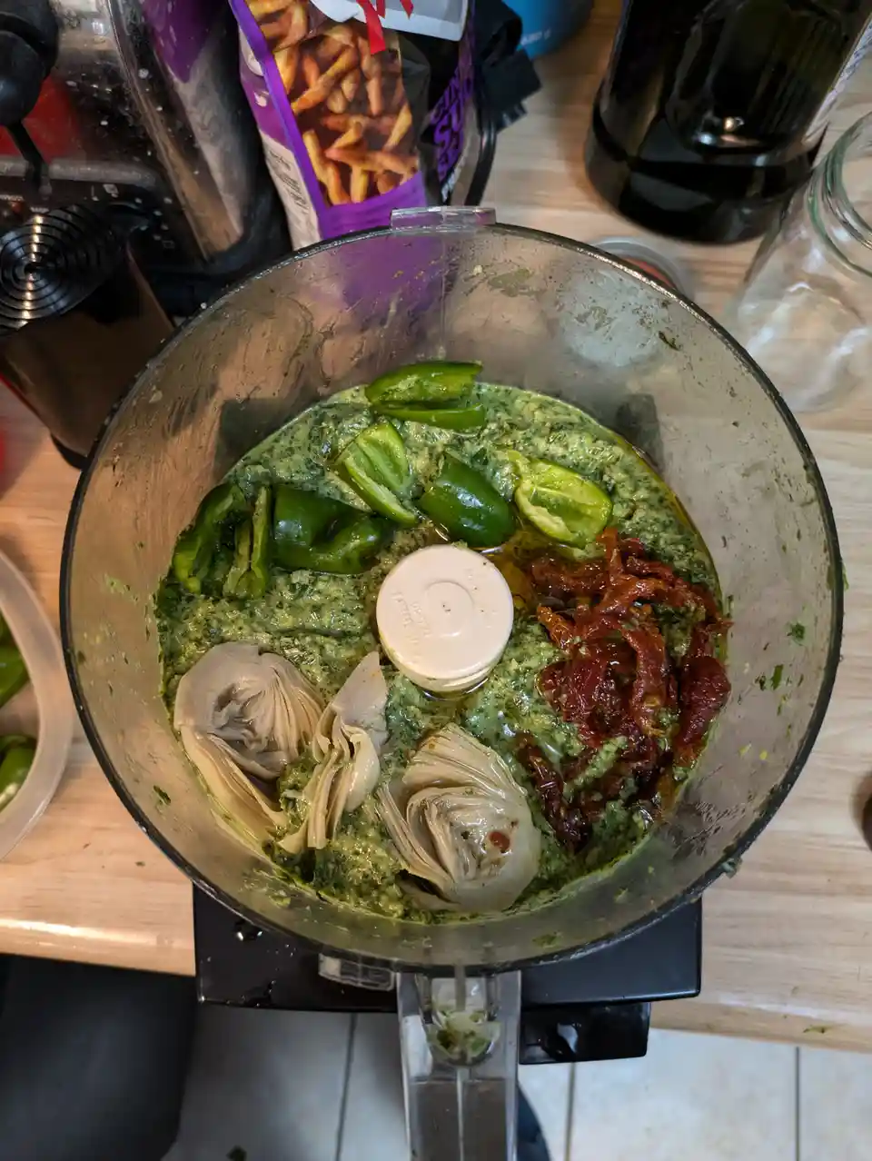A food processor bowl contains a green pesto base with whole green peppers, artichoke hearts, and sun-dried tomatoes added…
