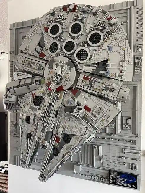 A large, intricate LEGO model of the Millennium Falcon spaceship is displayed on a gray LEGO-built wall, viewed from above…