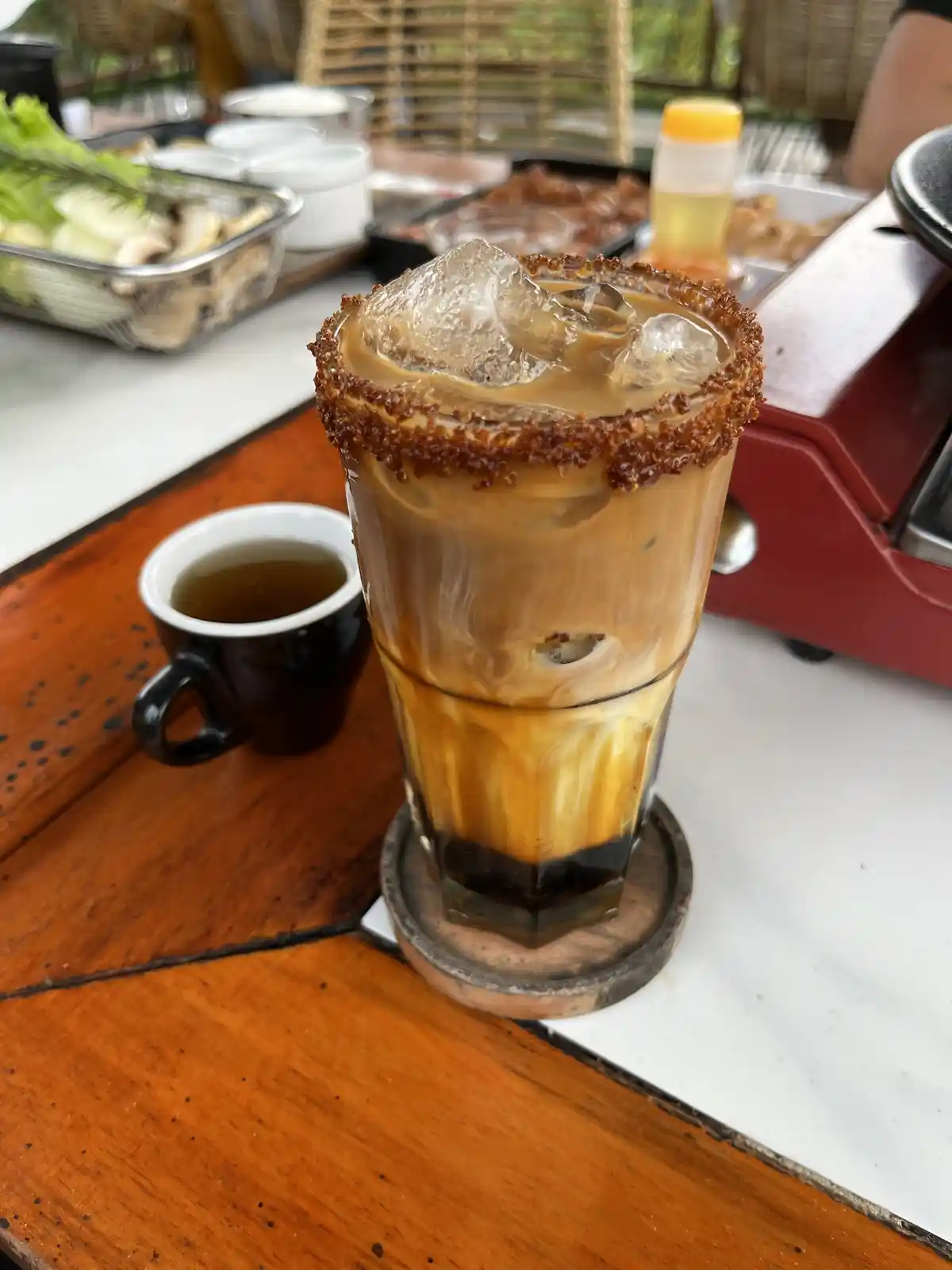 A tall glass of iced coffee with a caramelized sugar rim and large ice cubes sits on a wooden coaster on a table, next to a…