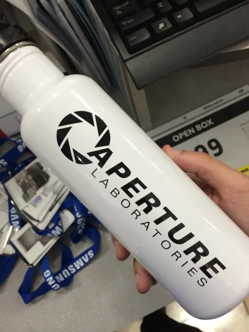 A hand holds a white water bottle with the black 'Aperture Laboratories' logo and text, featuring a camera aperture symbol…