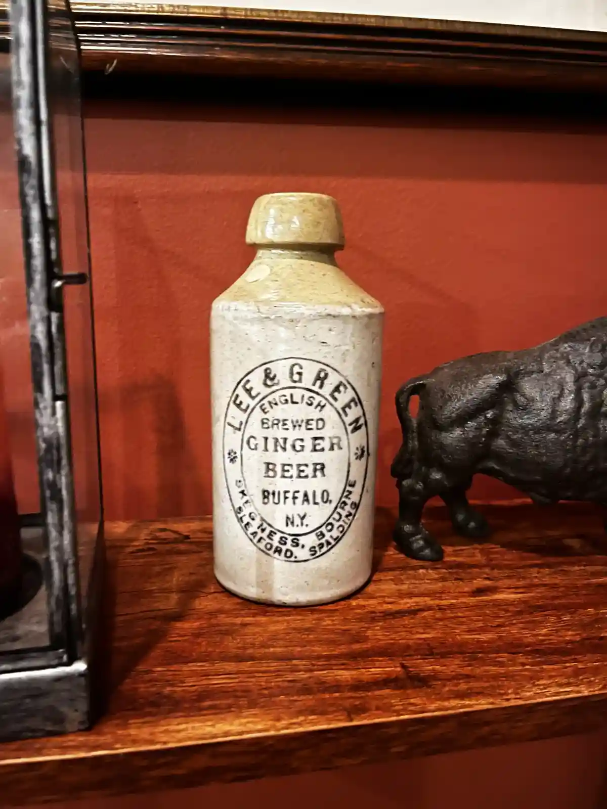 A vintage ceramic ginger beer bottle labeled 'Lee & Green English Brewed Ginger Beer Buffalo, N.Y.' sits on a wooden shelf…