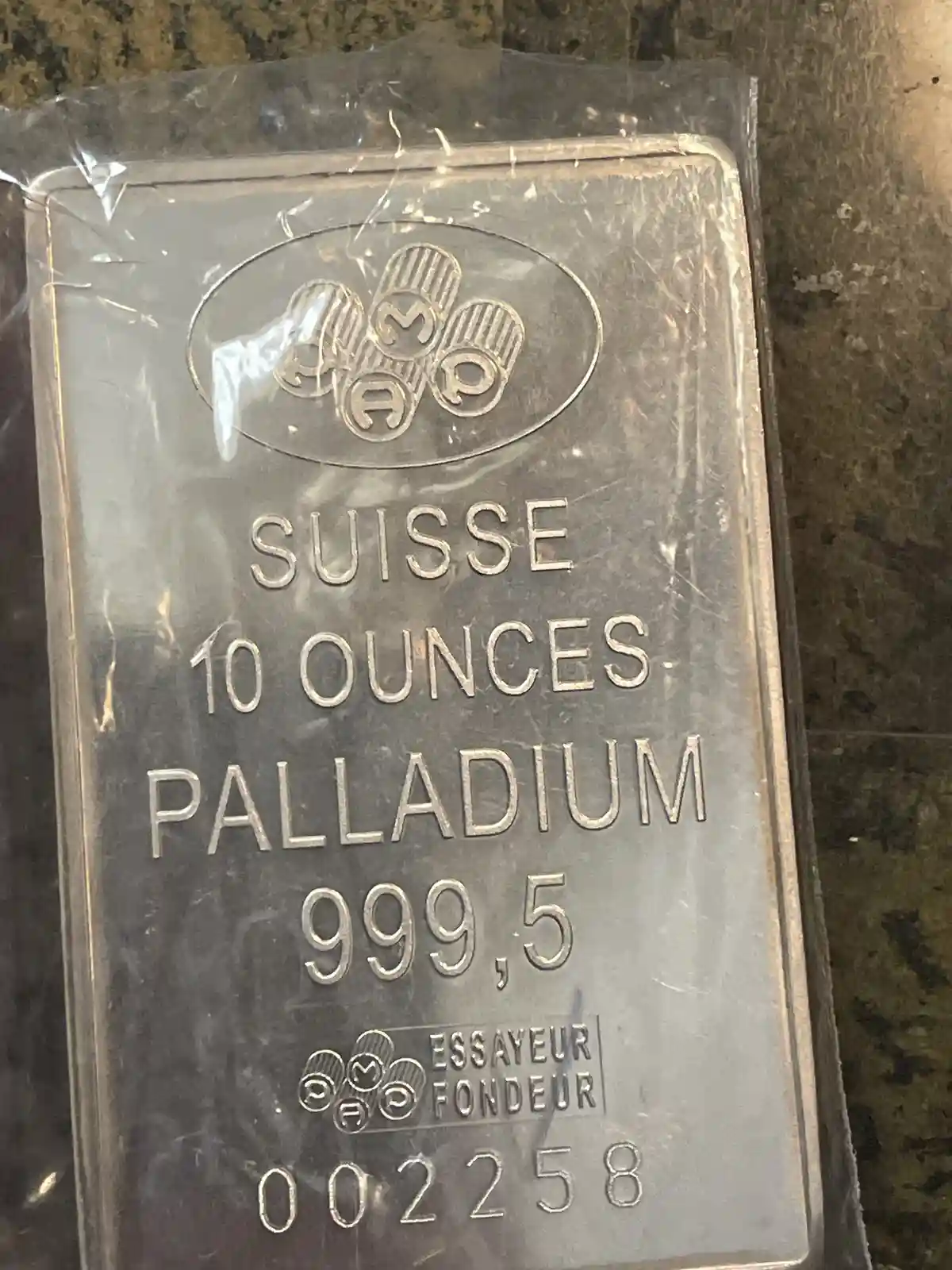 A close-up photo of a rectangular, silver-colored metal bar sealed in clear plastic, resting on a speckled countertop. The…