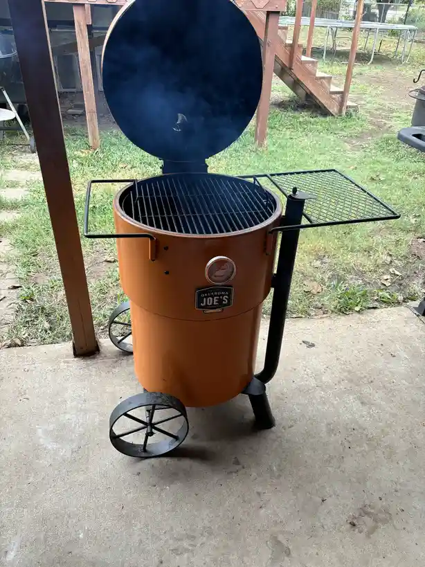 A large, orange, cylindrical Oklahoma Joe's smoker sits on a concrete patio with its lid open, emitting smoke. The smoker…