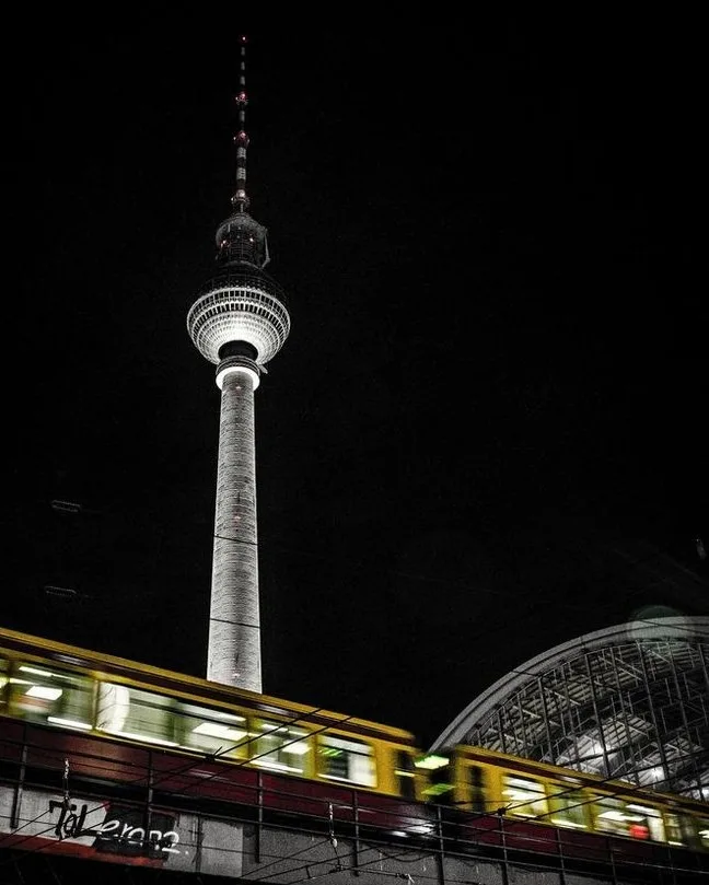 A nighttime photo of the Berlin TV Tower, with a yellow train blurred in motion in the foreground. The text 'Tal 2002' is…
