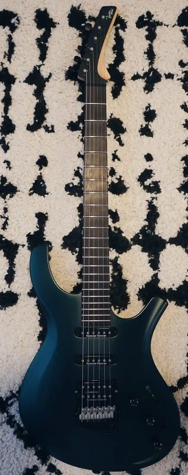 A dark teal electric guitar with a black fretboard and 'Seymour Duncan' pickups lies on a black-and-white patterned shag…