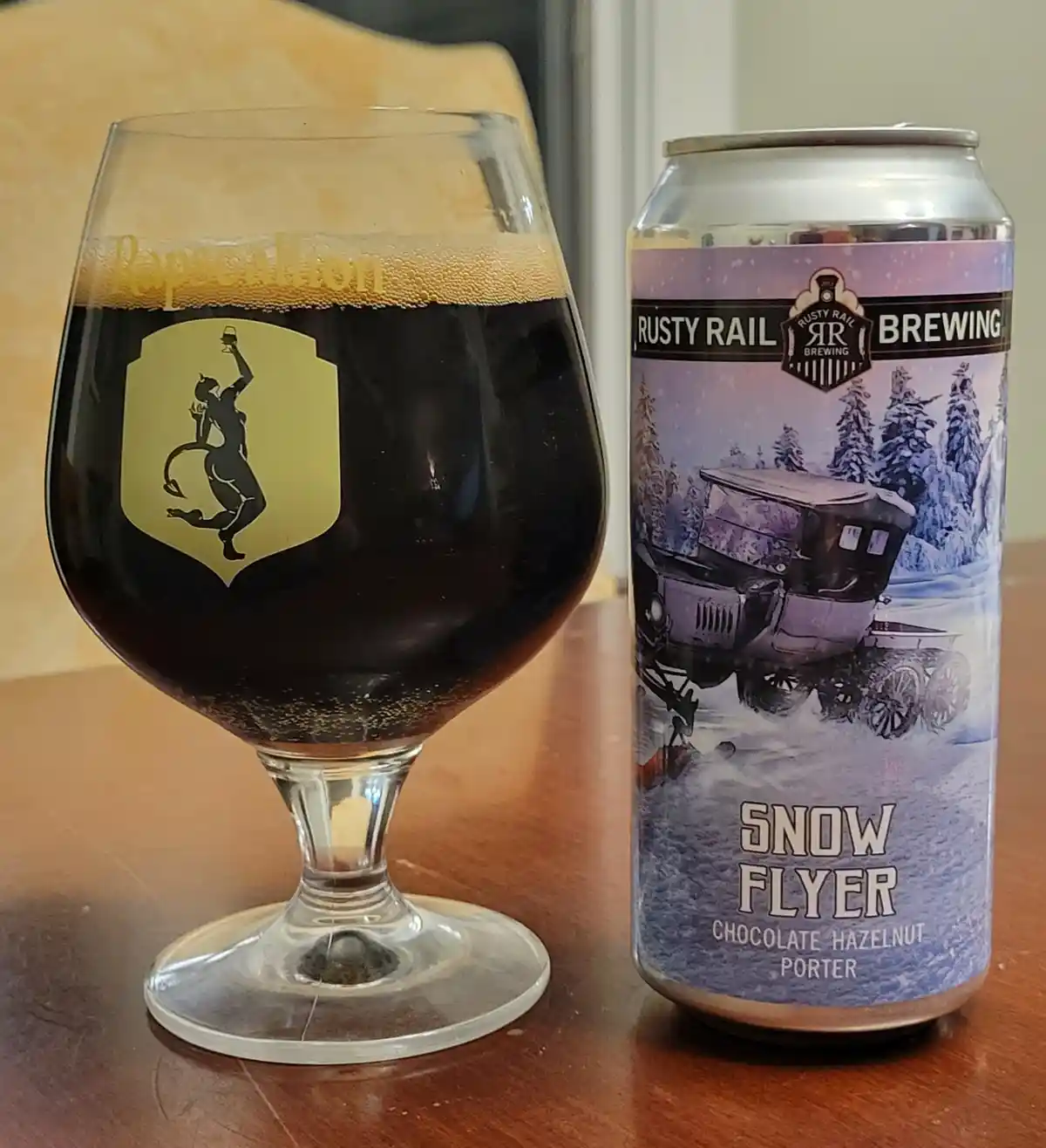 A dark beer in a stemmed glass with a gold 'Rapscallion' logo and devil silhouette sits next to a can of 'Rusty Rail…