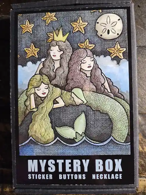 The image shows a product box for a 'Mystery Box' containing stickers, buttons, and a necklace. The box art features three…