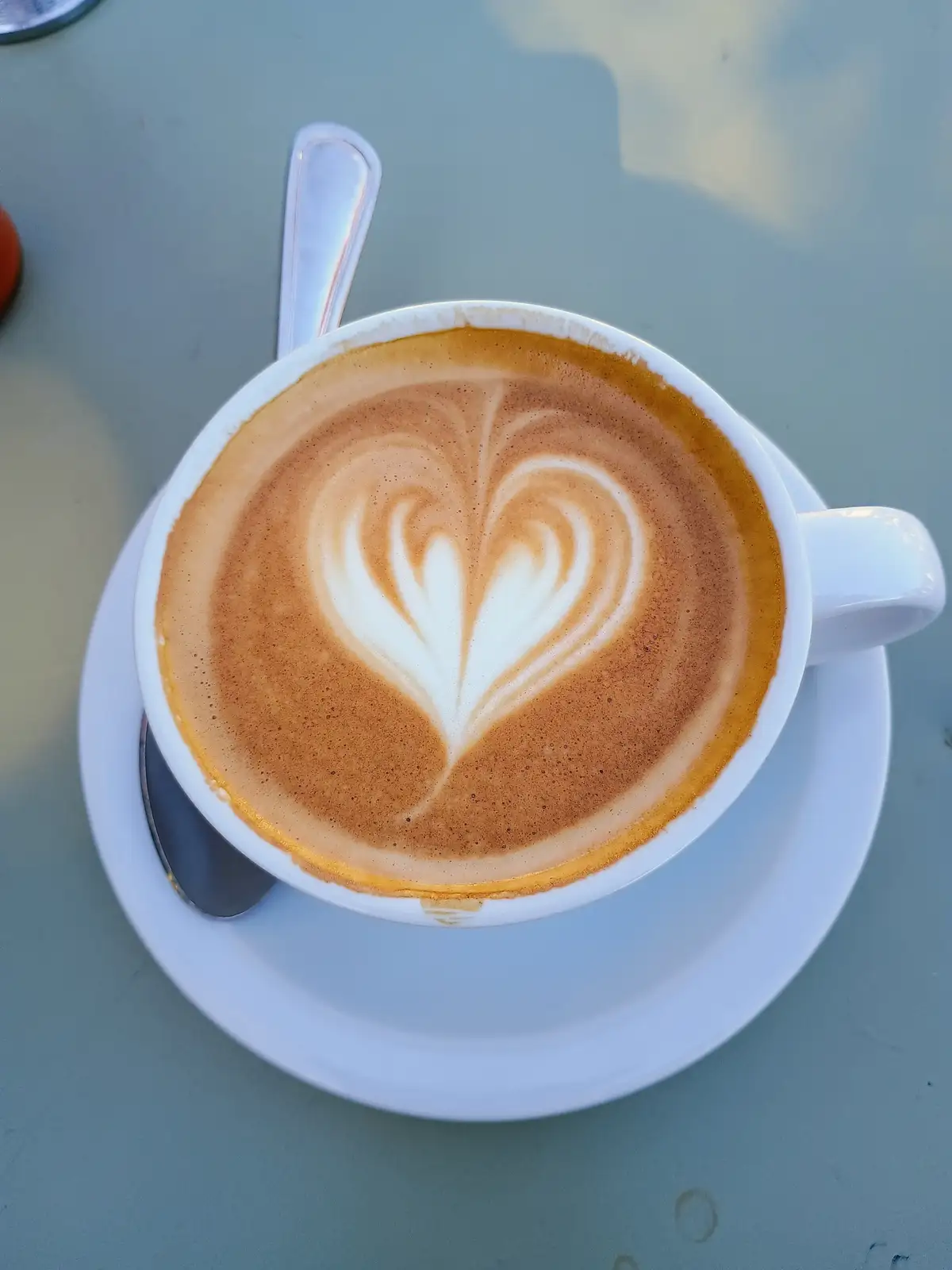 A top-down photo of a white ceramic coffee cup on a saucer, containing a latte with intricate heart-shaped latte art made…