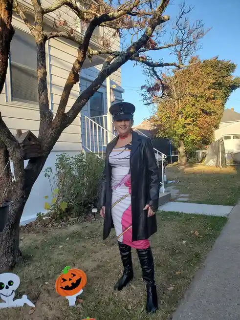 A woman in a Halloween costume stands in a yard with autumn trees and a house behind her. She wears a black police-style…