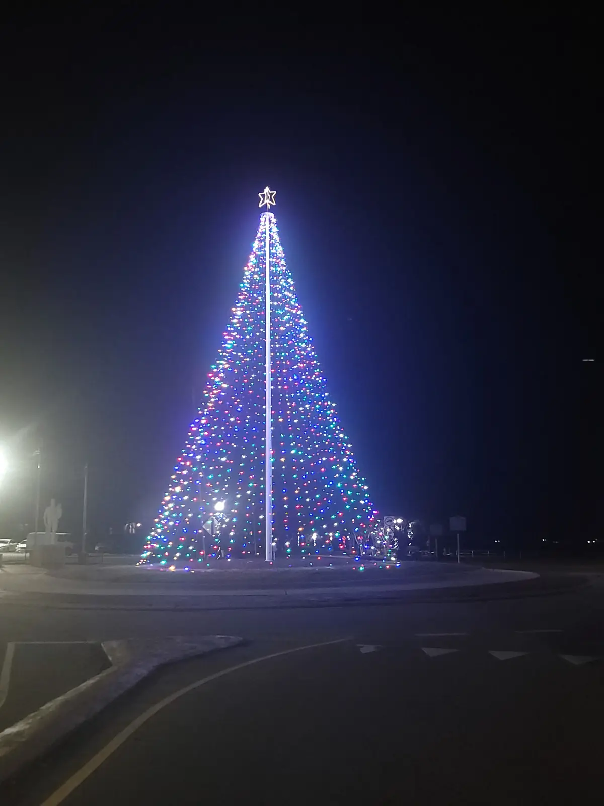A tall, brightly lit Christmas tree adorned with multicolored lights and topped with a star stands at night in a public…