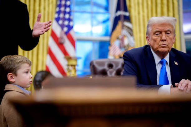 A young boy stands in front of a desk, looking up at a man in a suit seated behind it. The man, identified as Donald Trump…
