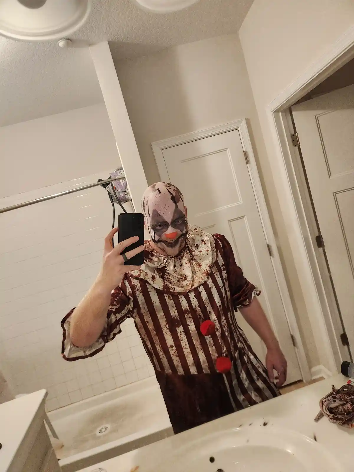 A person in a horror-themed clown costume with smeared makeup and blood-like stains on their face and striped shirt takes a…