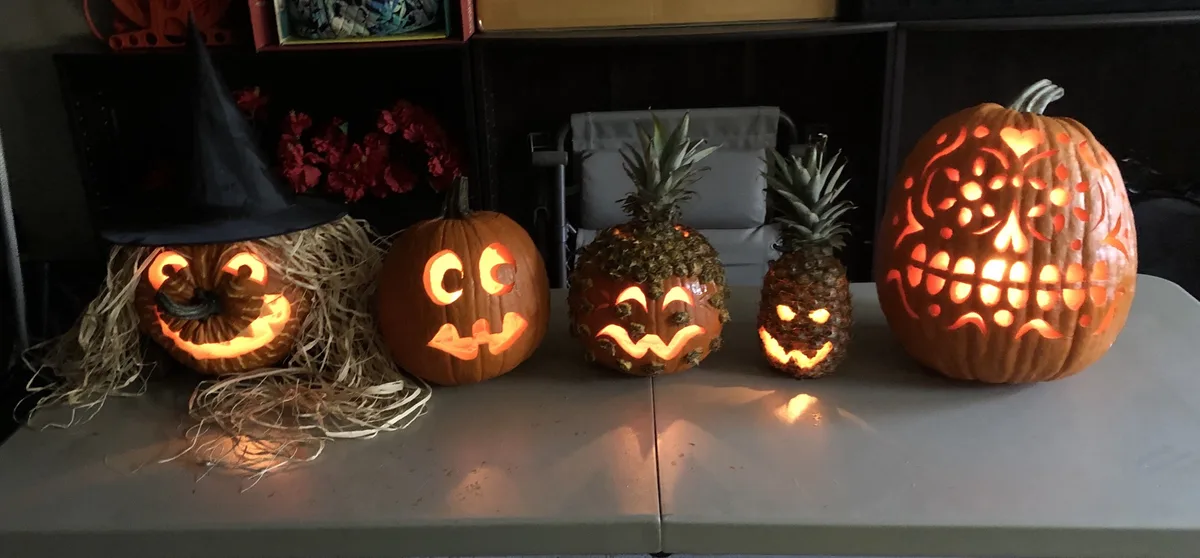 Five carved Halloween decorations sit on a table, including pumpkins and pineapples with glowing faces, one pumpkin wears a…