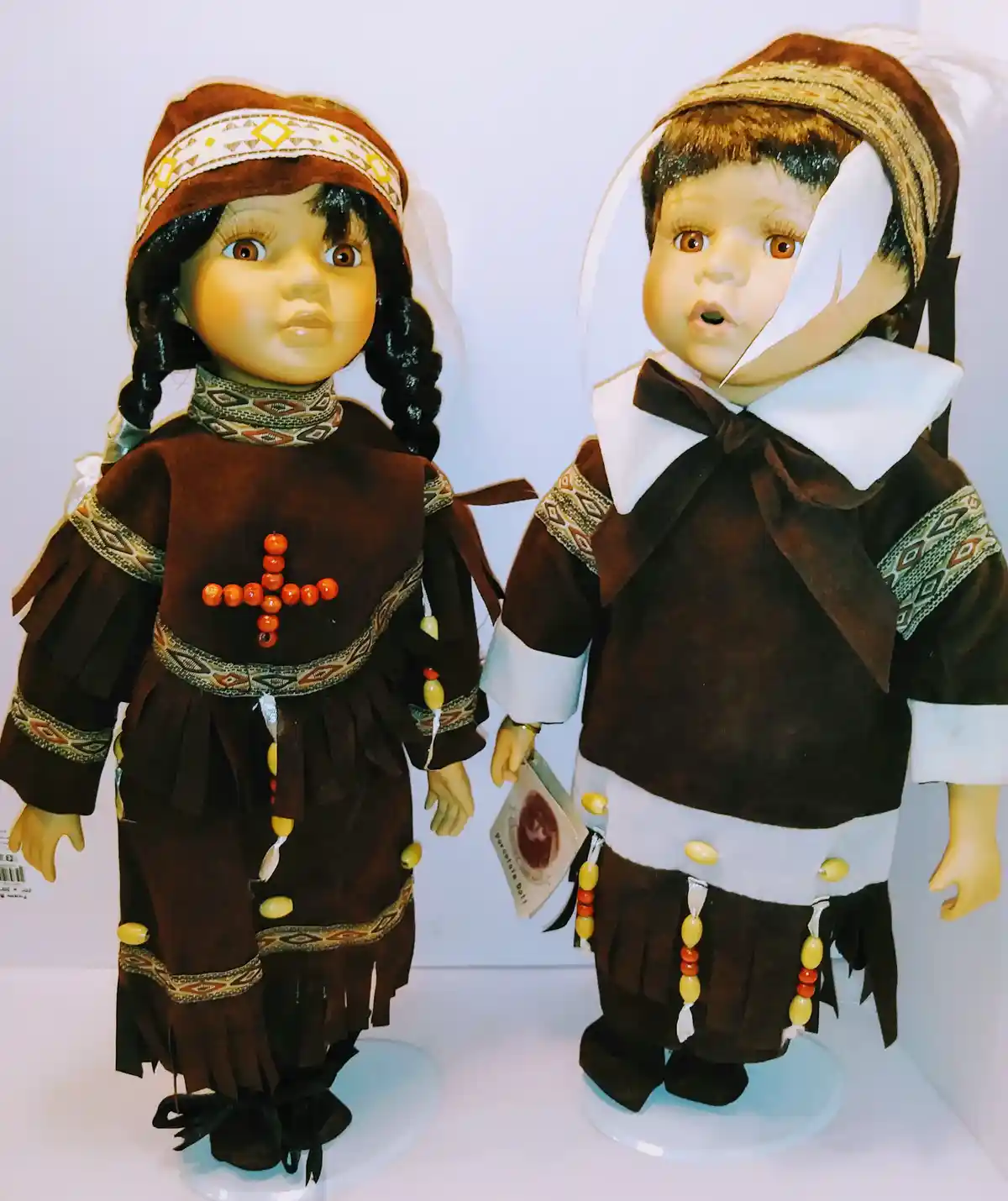 Two porcelain dolls dressed in traditional Native American and Pilgrim attire stand side by side, both with wide-eyed…