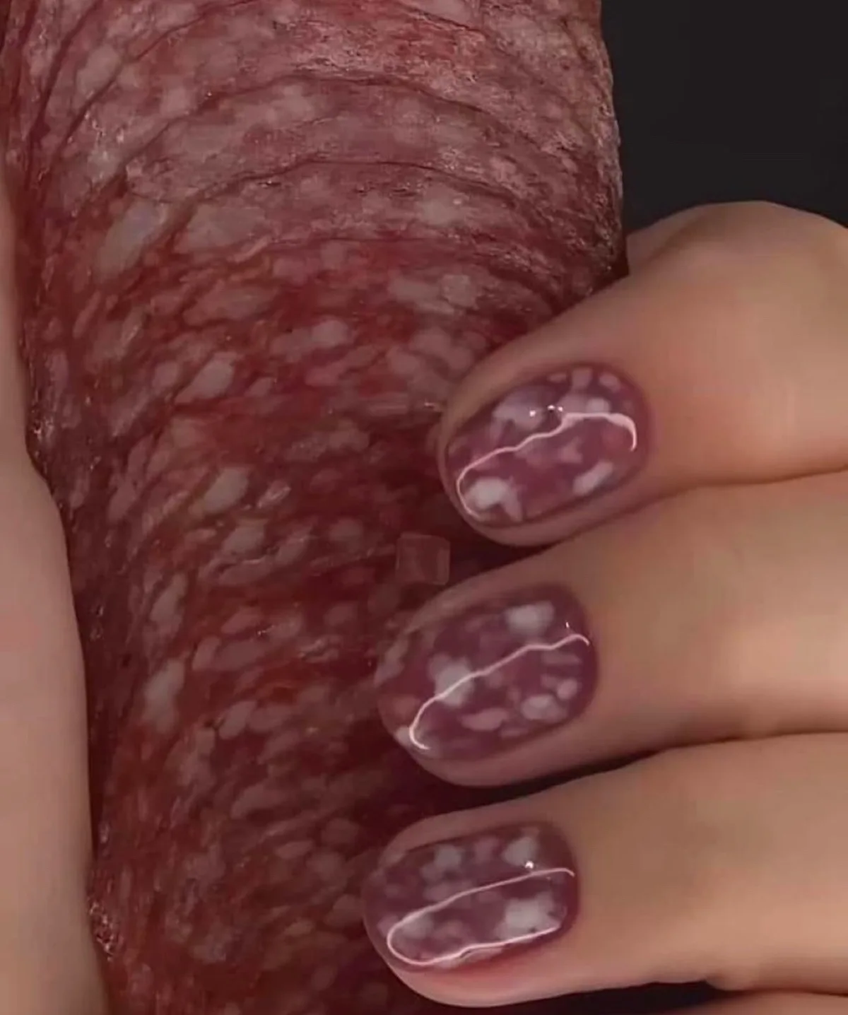 A close-up image shows a hand with fingernails painted to resemble salami, featuring a marbled red and white pattern with a…