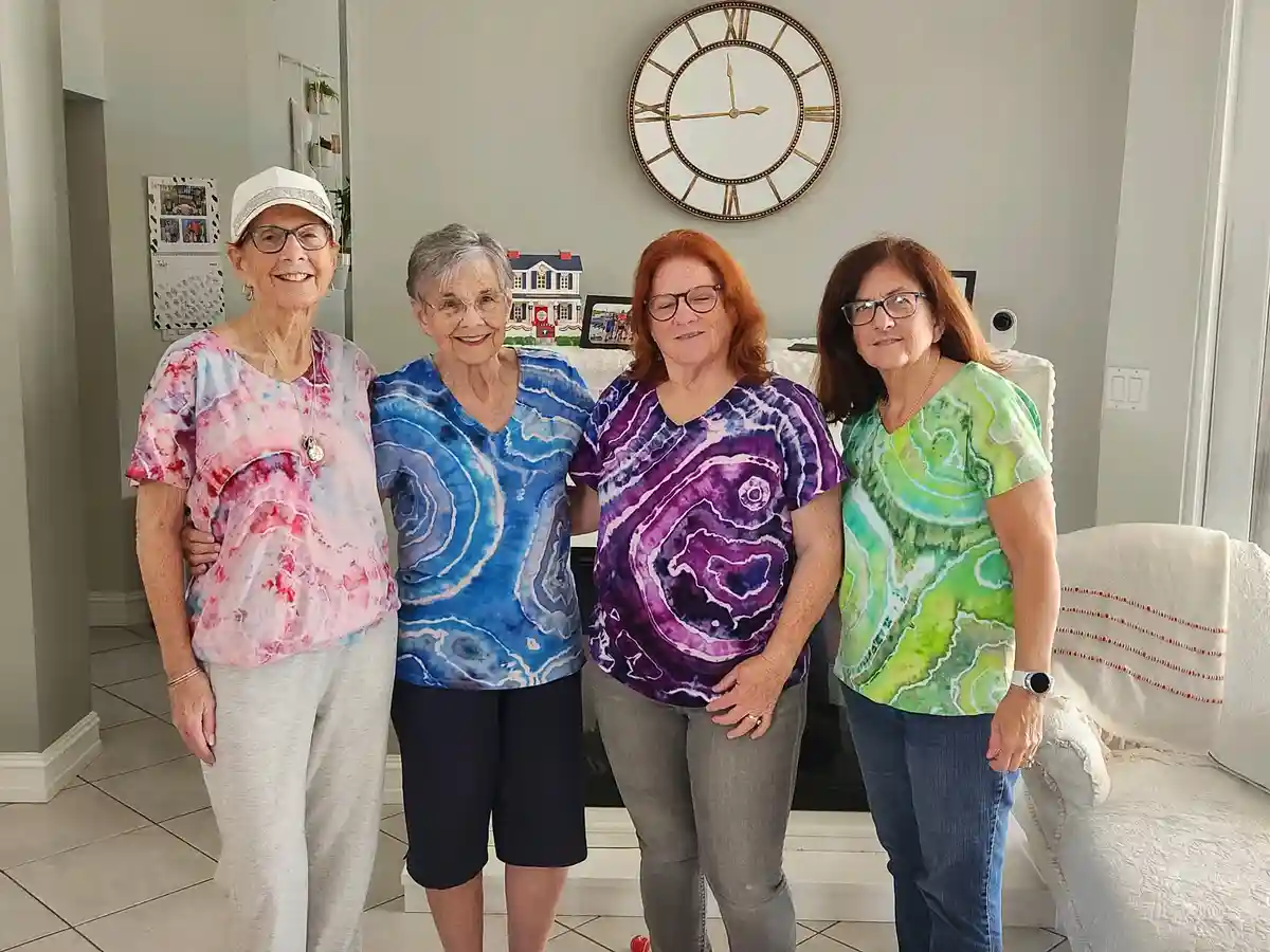 four smiling older women stand together in a living room, each wearing a brightly colored, patterned shirt resembling agate…