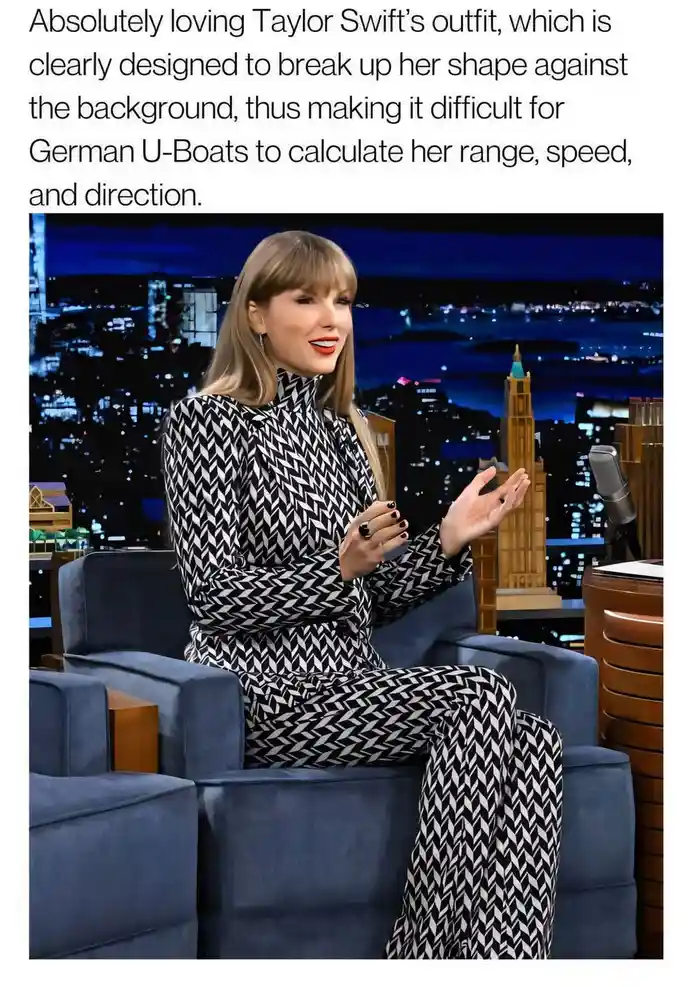 Taylor Swift is seated in a blue armchair on a talk show set with a nighttime city skyline backdrop. She wears a…