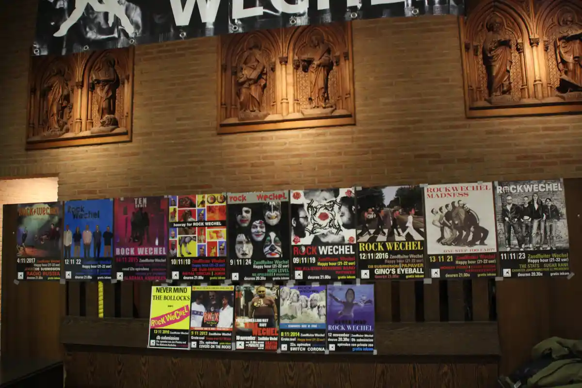 A row of colorful concert posters for 'Rock Wechel' events are taped to a wooden surface in front of a brick wall adorned…
