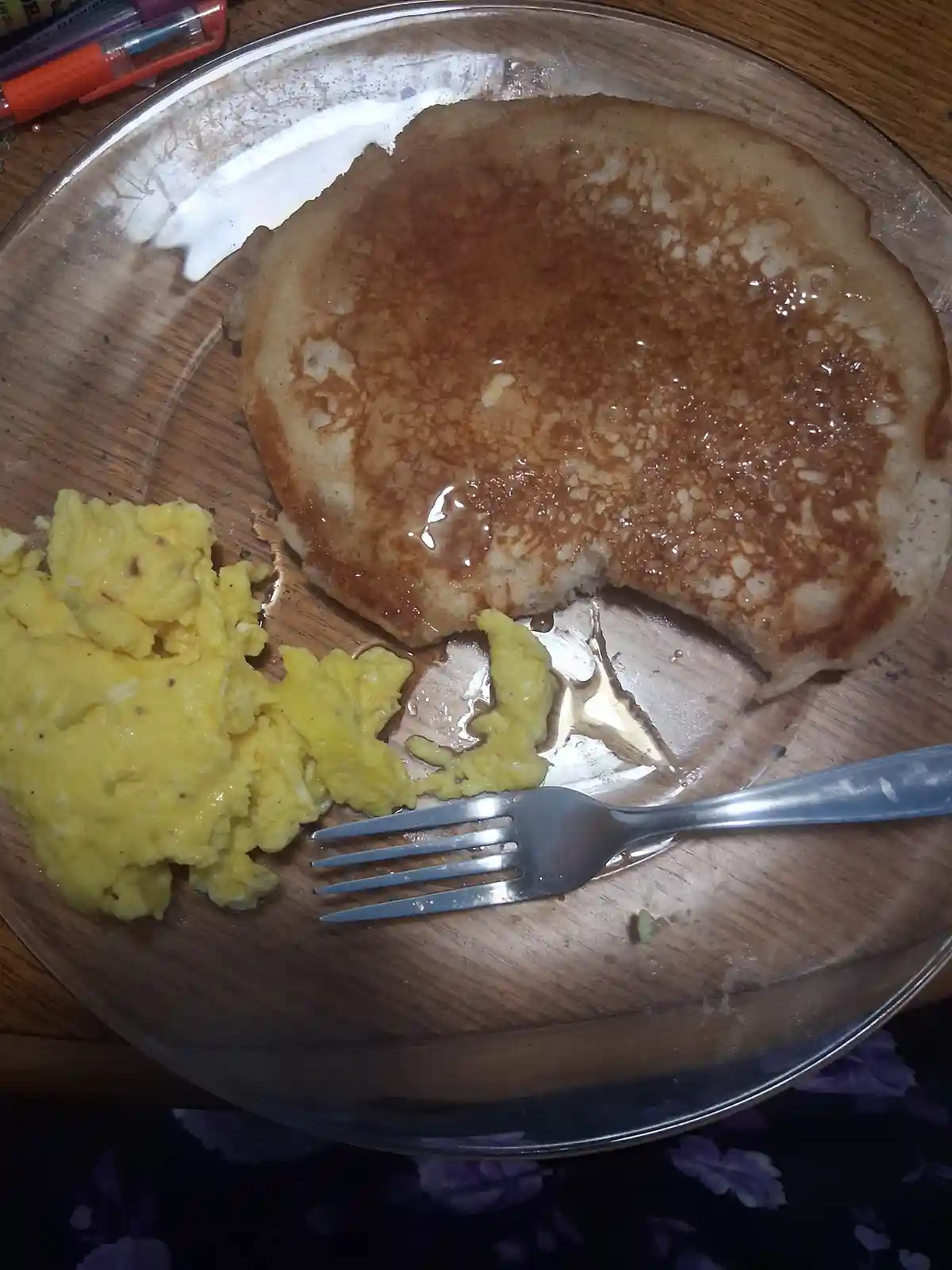 A close-up photo shows a partially eaten pancake with syrup and a side of scrambled eggs on a clear glass plate, with a fork…
