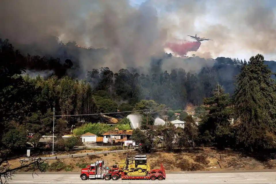 A red fire truck hauling a yellow bulldozer on a flatbed trailer drives on a highway, while in the background, a wildfire…