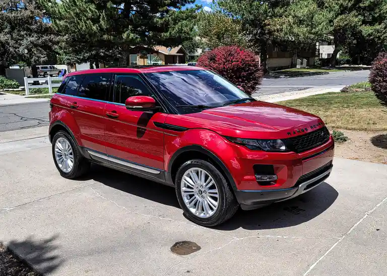 A shiny red Range Rover Evoque is parked on a cracked concrete driveway in a suburban neighborhood with trees and houses in…