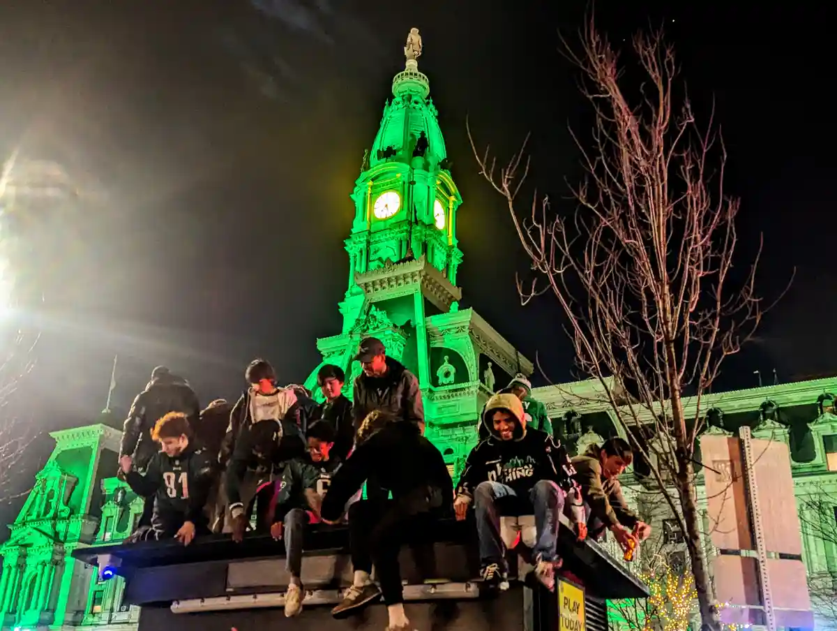 A group of young people are sitting and standing on the top of a large public structure at night, with a grand, ornate clock…