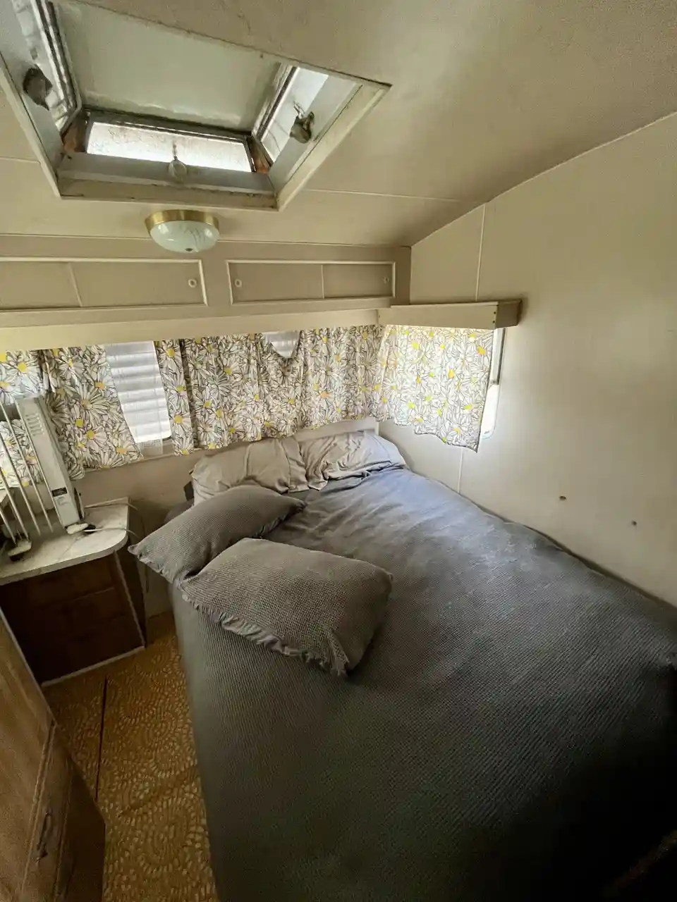 An interior shot of a vintage camper or trailer bedroom with a bed, daisy-patterned curtains, a small desk, and a skylight…