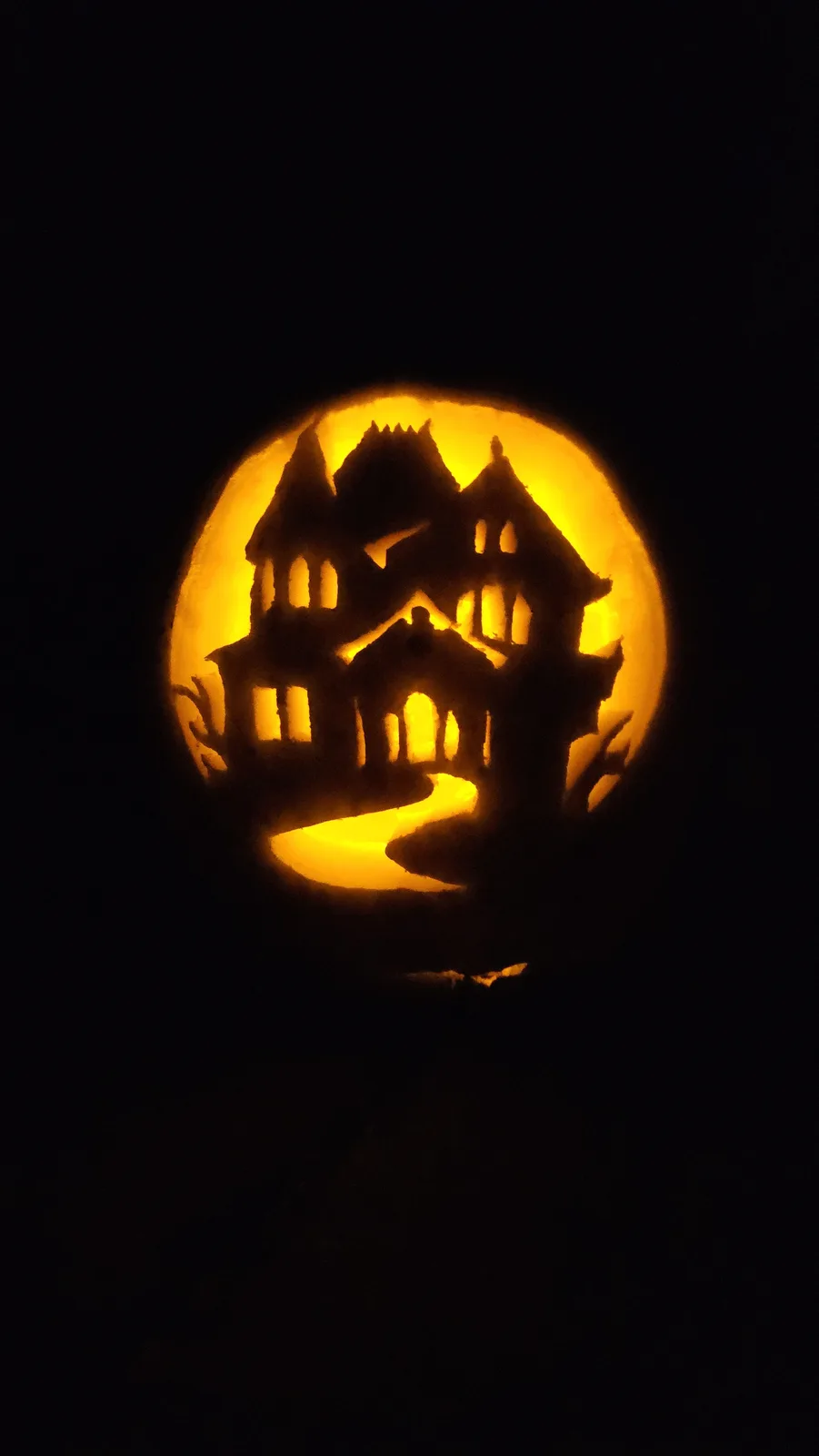 A carved pumpkin illuminated from within, displaying a detailed silhouette of a spooky, multi-story haunted house with…