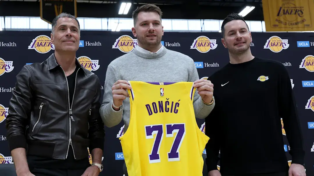 Three men stand in front of a Los Angeles Lakers branded backdrop. The man in the center holds up a yellow Lakers jersey…
