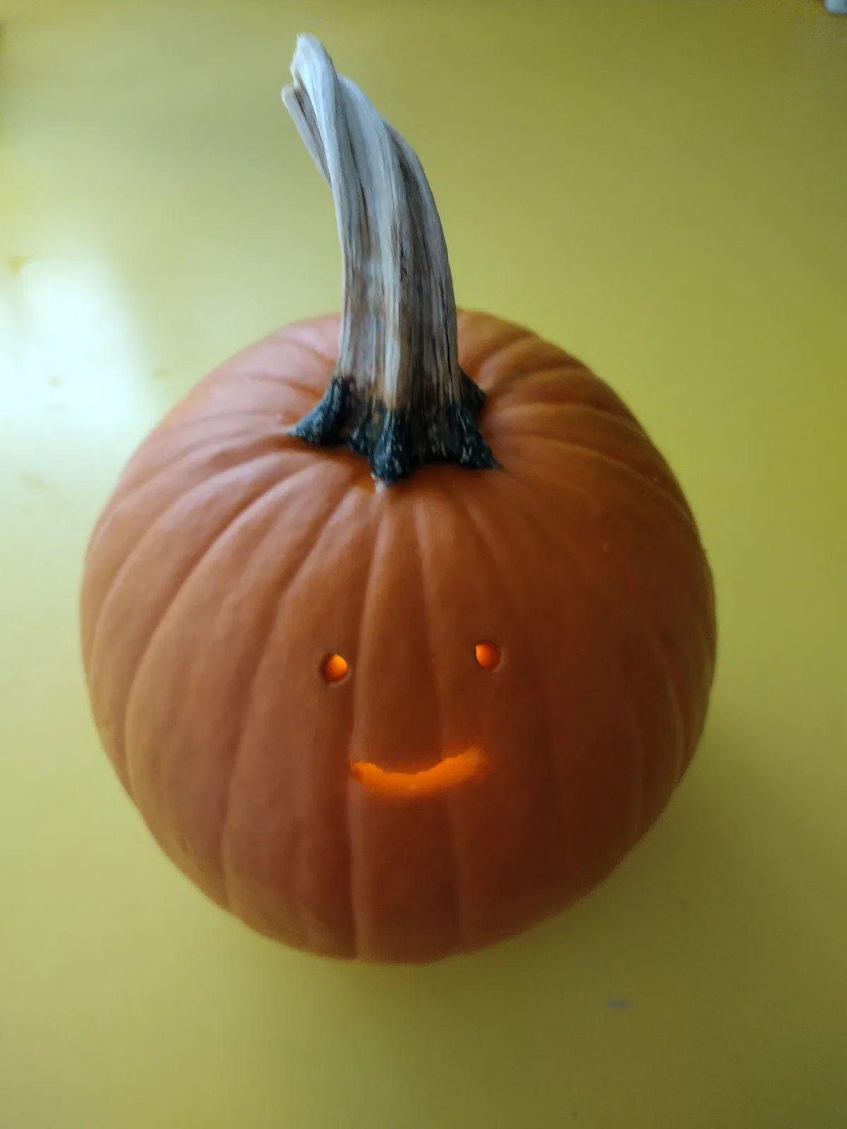 A close-up photo shows a small orange pumpkin with a carved smiling face, featuring two round eye holes and a curved mouth…