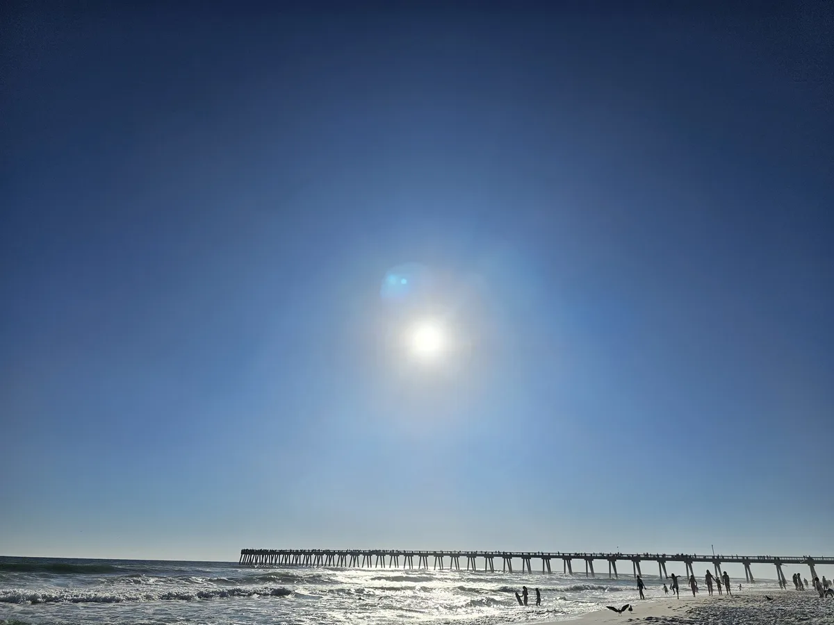 A bright, clear blue sky with the sun shining intensely, casting lens flares, above a long pier extending into the ocean…