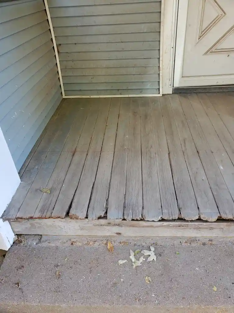 A close-up photo shows a weathered wooden porch floor with visibly warped and broken planks, leading up to a white door with…