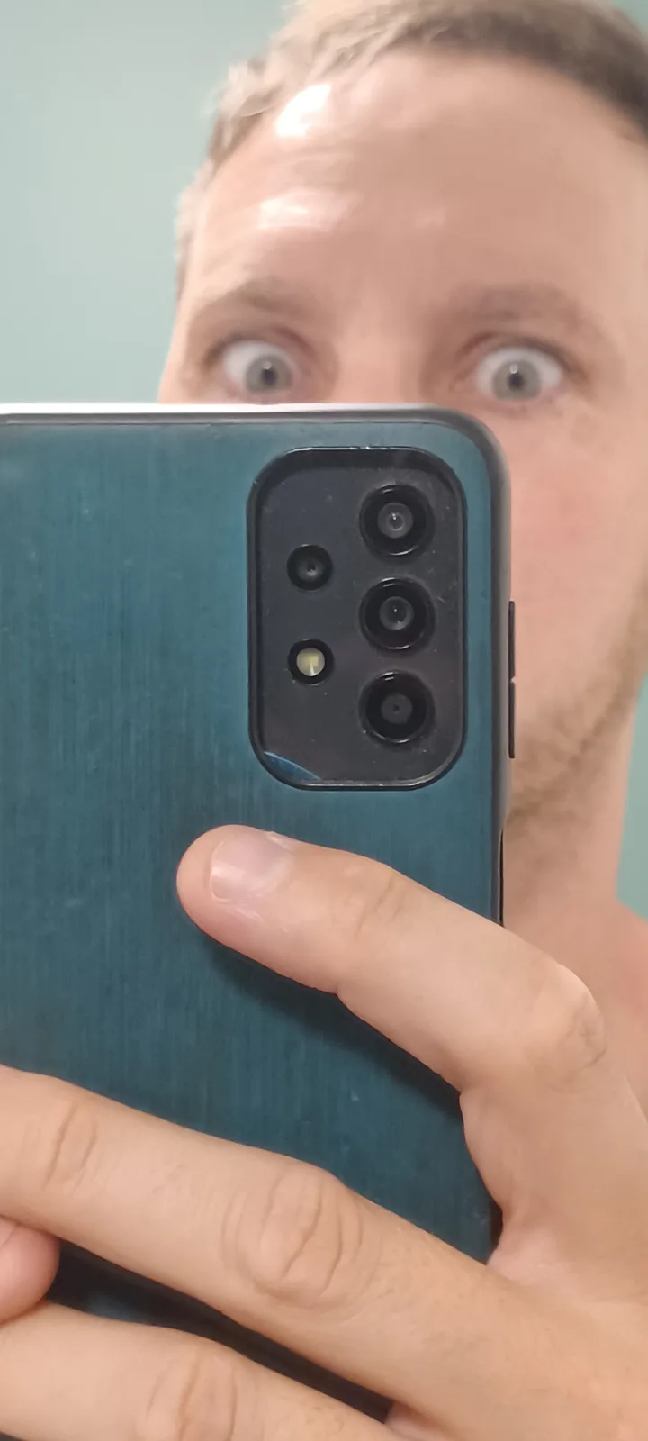 A person holds a teal smartphone with a visible rear camera module in front of their face, obscuring most of it, while their…