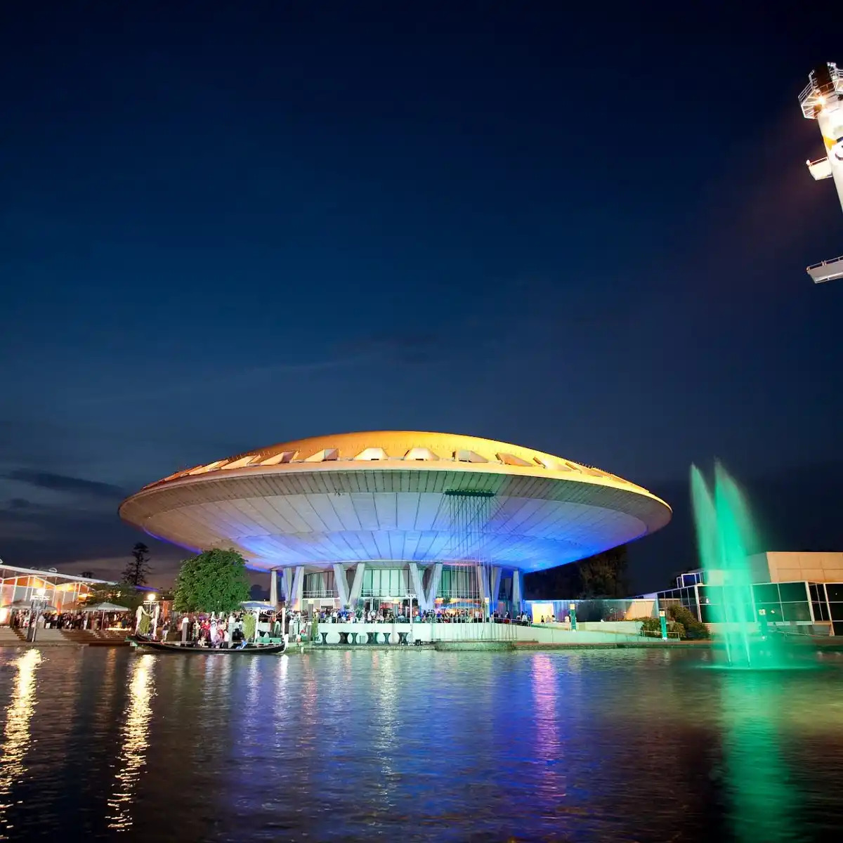 A nighttime photo of a large, saucer-shaped building illuminated with blue and gold lights, situated on the edge of a body…