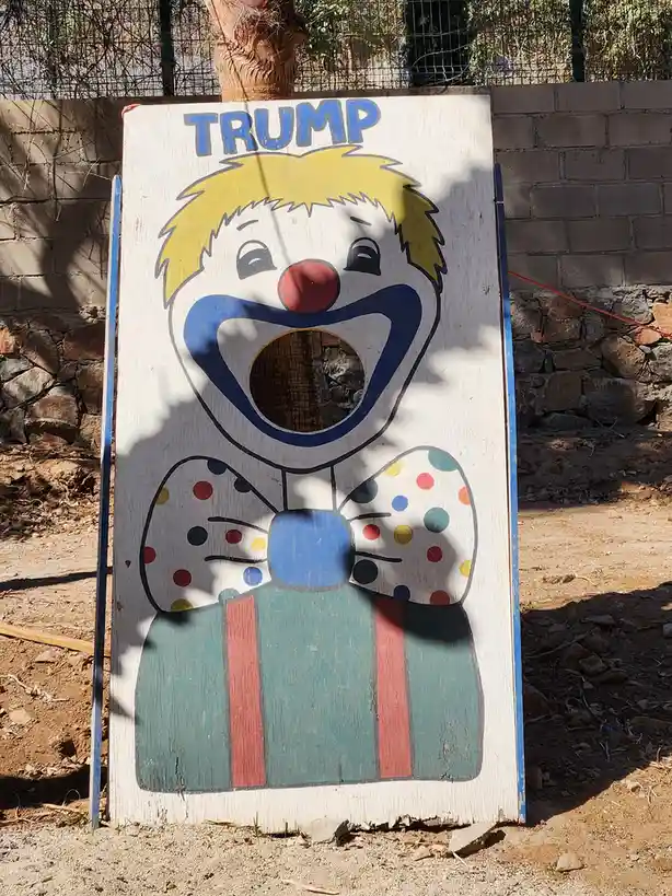 A weathered wooden cutout of a clown with a large painted smile and a hole in its mouth stands outdoors. The clown has…
