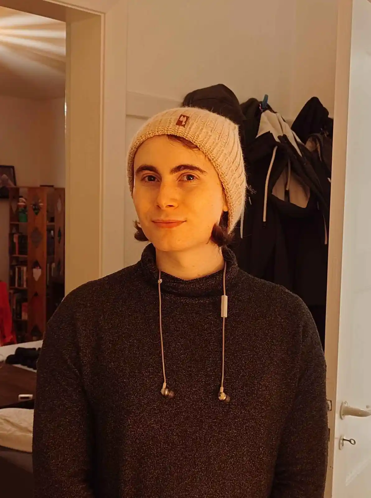 A person with shoulder-length brown hair wears a beige knit beanie and a dark grey turtleneck sweater, with white earbuds…